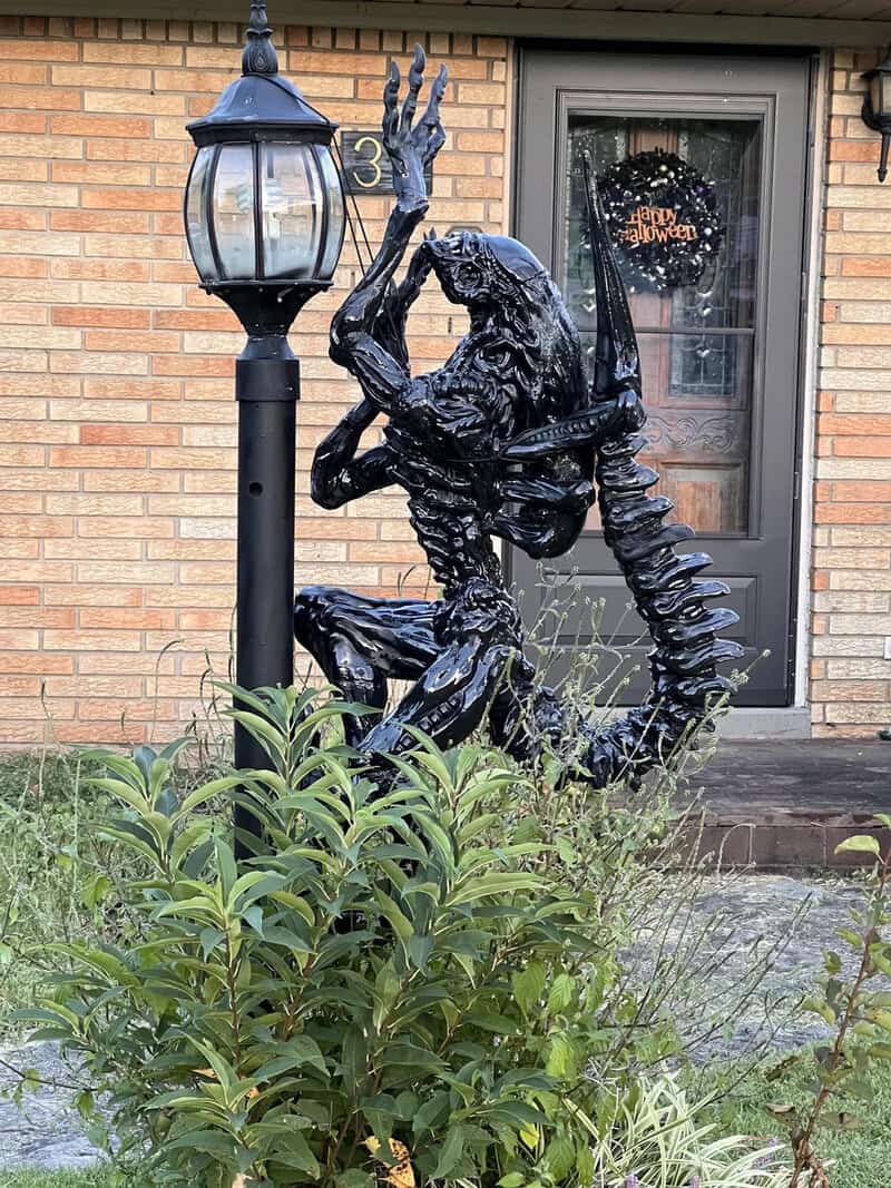 Black alien-like sculpture crouching beside a lamppost in front of a house with Halloween wreath on.