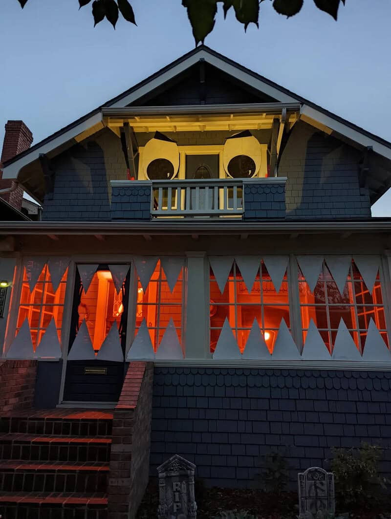 Haunted house with orange triangle bunting across porch, Halloween lights glowing inside windows, a}.
