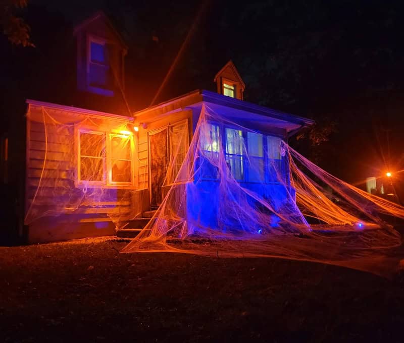 House at night wrapped in orange and blue Halloween spiderweb lights, spooky porch glow.