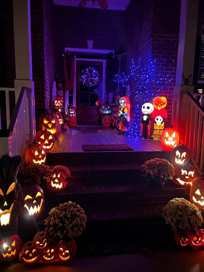 Halloween-decorated porch with carved pumpkins, blue string lights, skeletons and a saintly figure,.