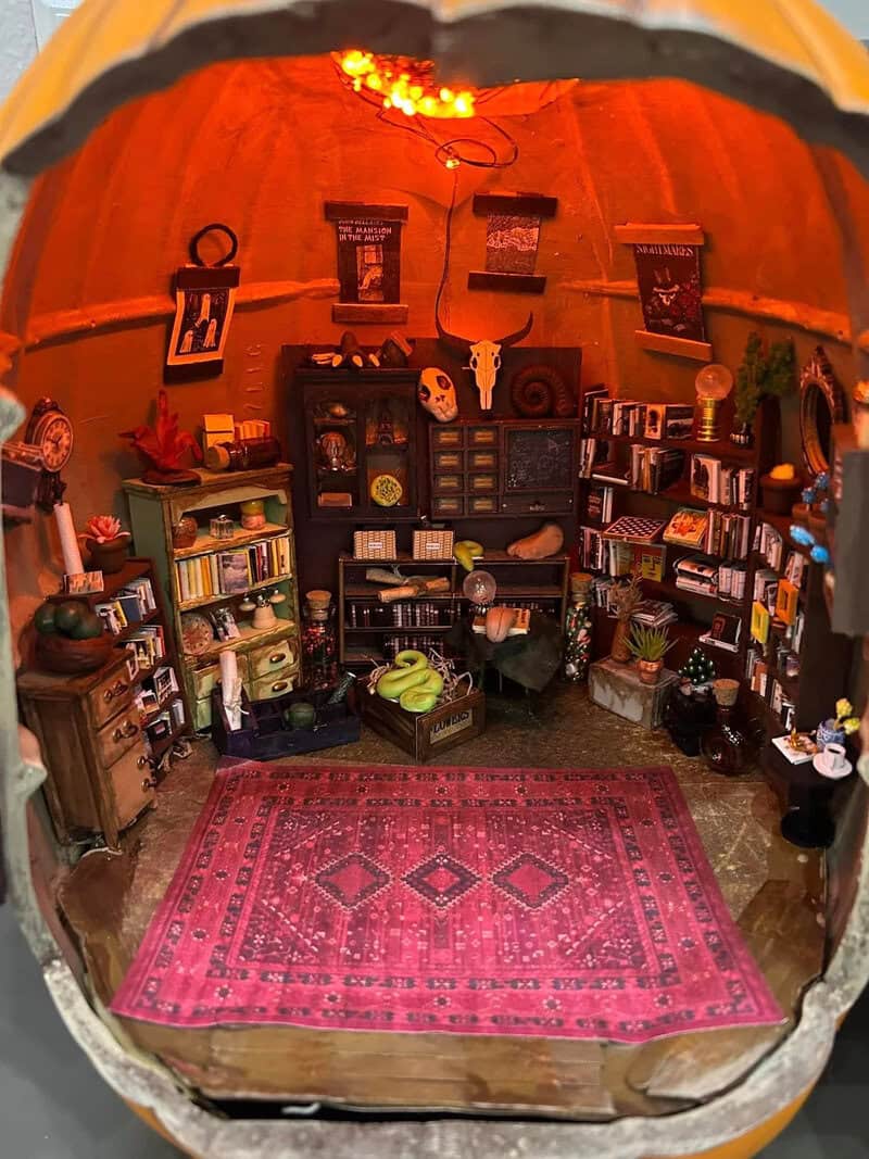 Cozy Halloween-themed dollhouse-like room with shelves of miniature items, pink patterned rug, warm灯.