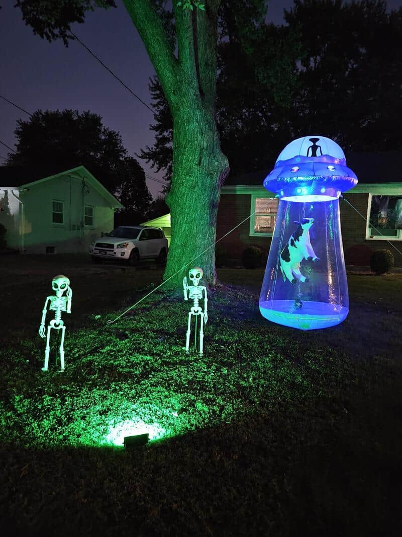 Halloween yard display with green-lit skeletons and a glowing UFO prop over a hanging ghost figure.