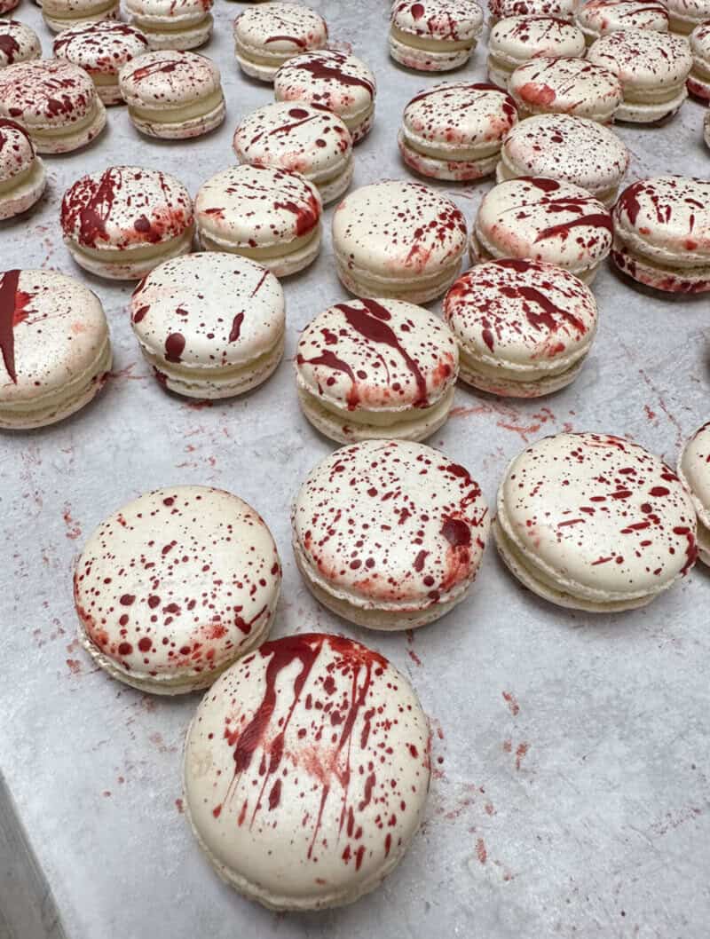 Macarons with white shells splattered with red filling on a baking sheet; many arranged in rows.