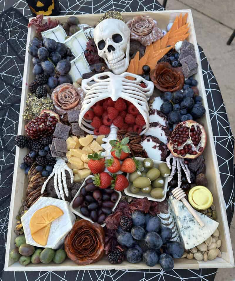 Halloween charcuterie board with a skeletal centerpiece, berries, cheese, grapes, olives, and autumn.