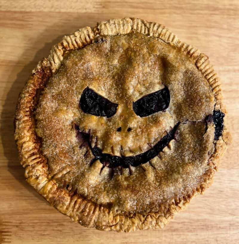 Smiling pumpkin pie with a mischievous carved face, cinnamon crust edges, on a wooden surface.