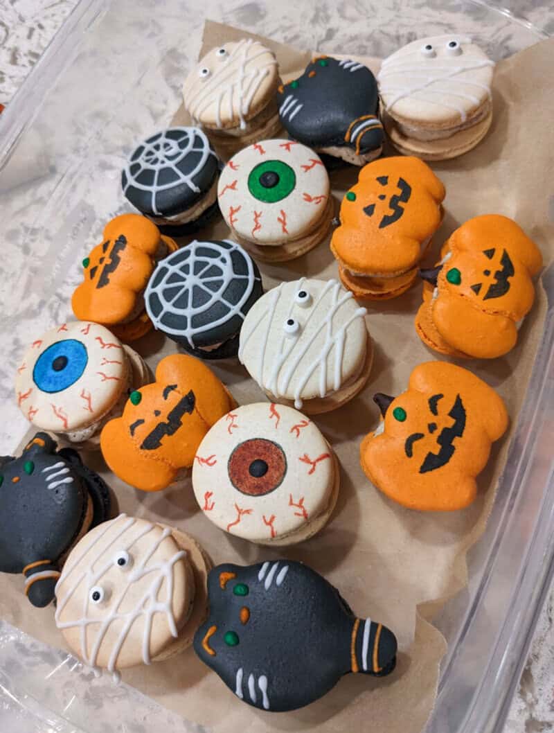Assorted Halloween macarons shaped as pumpkins, ghosts, eyeballs, and spider webs with orange, white.