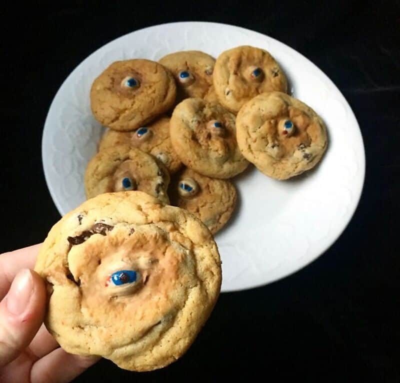 Hand holding a spooky eyeball cookie with chocolate center, others on plate in background.
