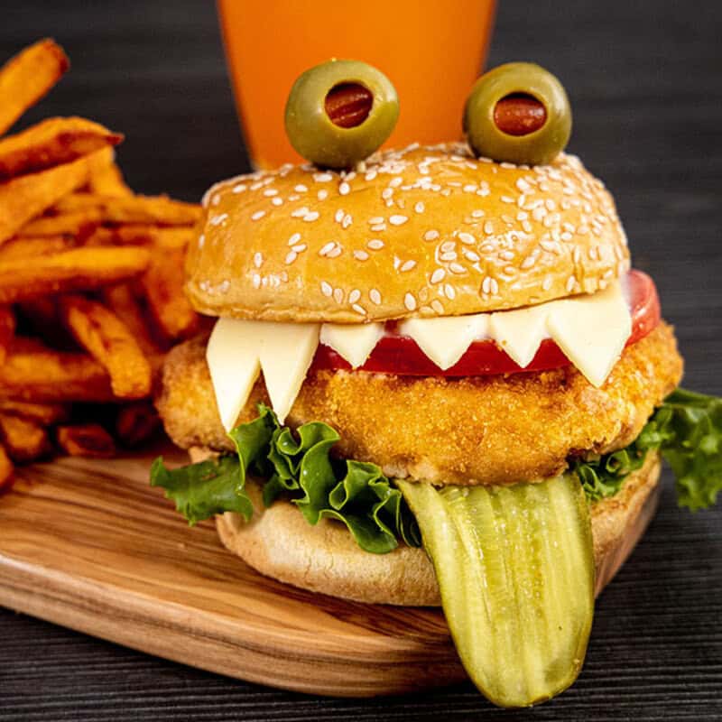 Halloween-themed burger with sesame bun, olives eyes, string cheese fangs, pickle tongue, lettuce, &.
