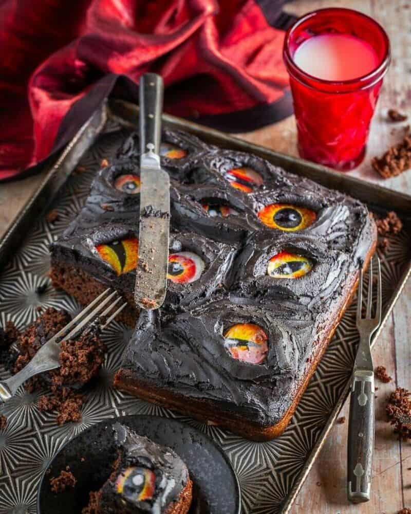 Dark chocolate cake with colored candy eyeballs on top, a red glass of milk, and kitchen utensils on.