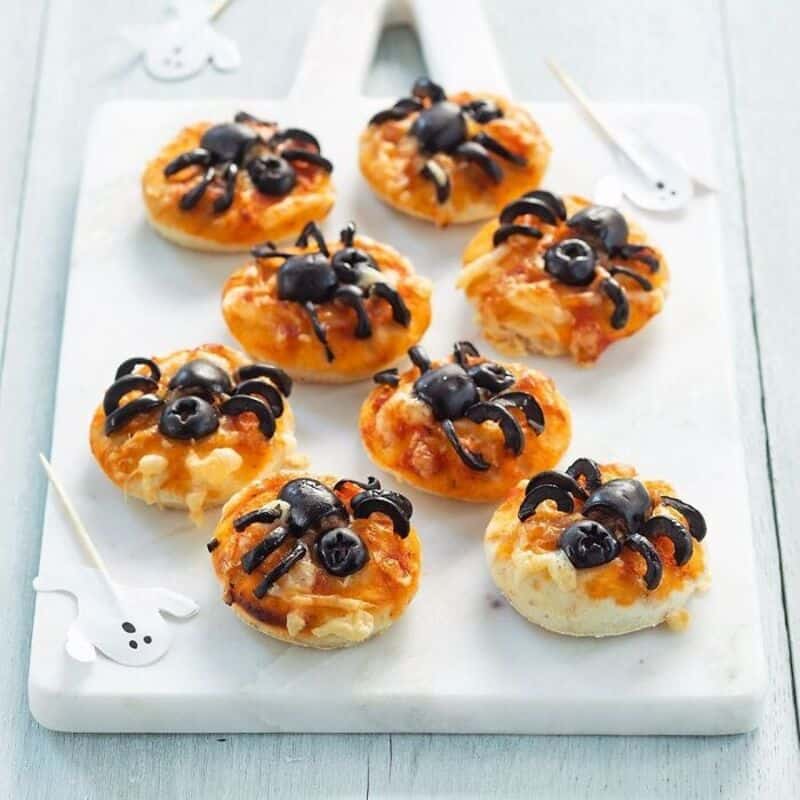 A tray of Halloween spider pastries with black olive bodies and legs on a light board.