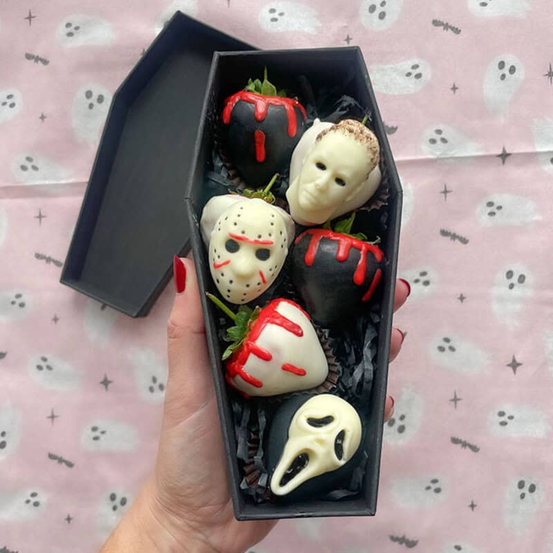 Halloween-themed chocolate strawberries shaped like masks and skulls in a coffin-shaped box.