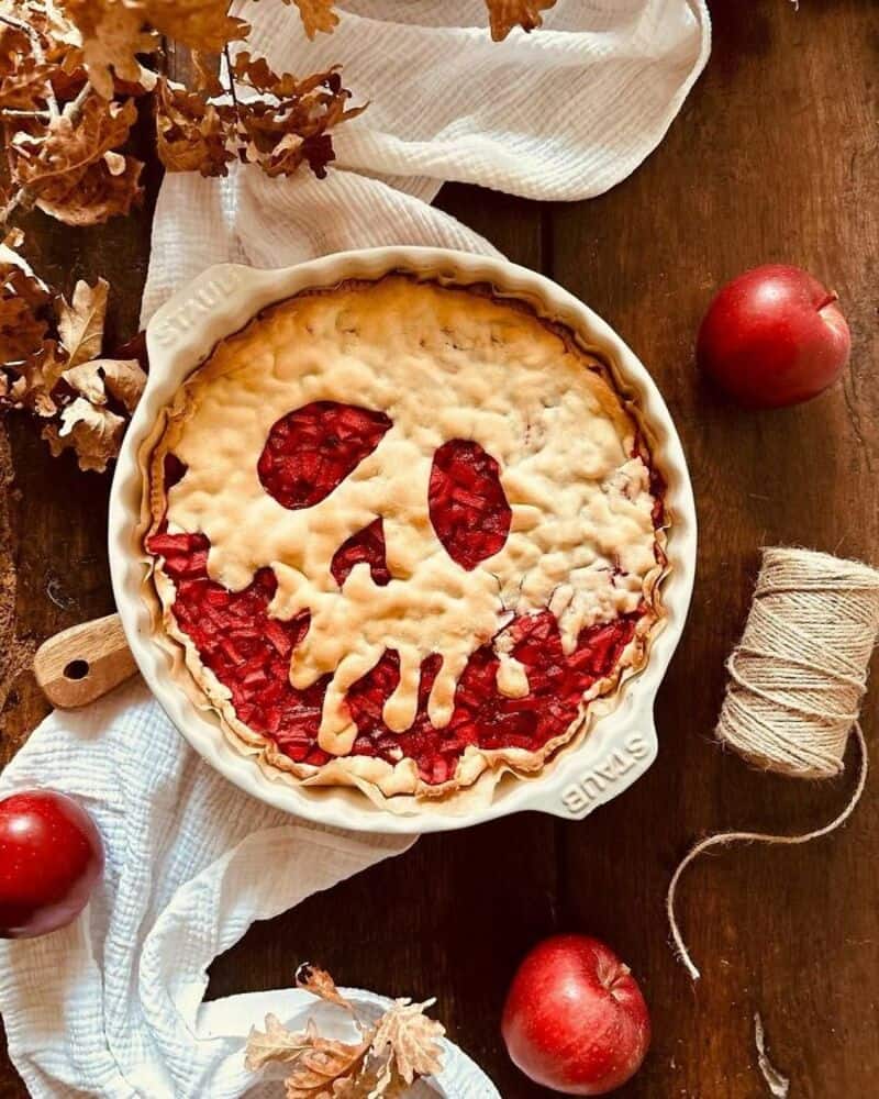 Pie with a skull face design on top, carved crust and red fruit filling in a round baking dish on a;.