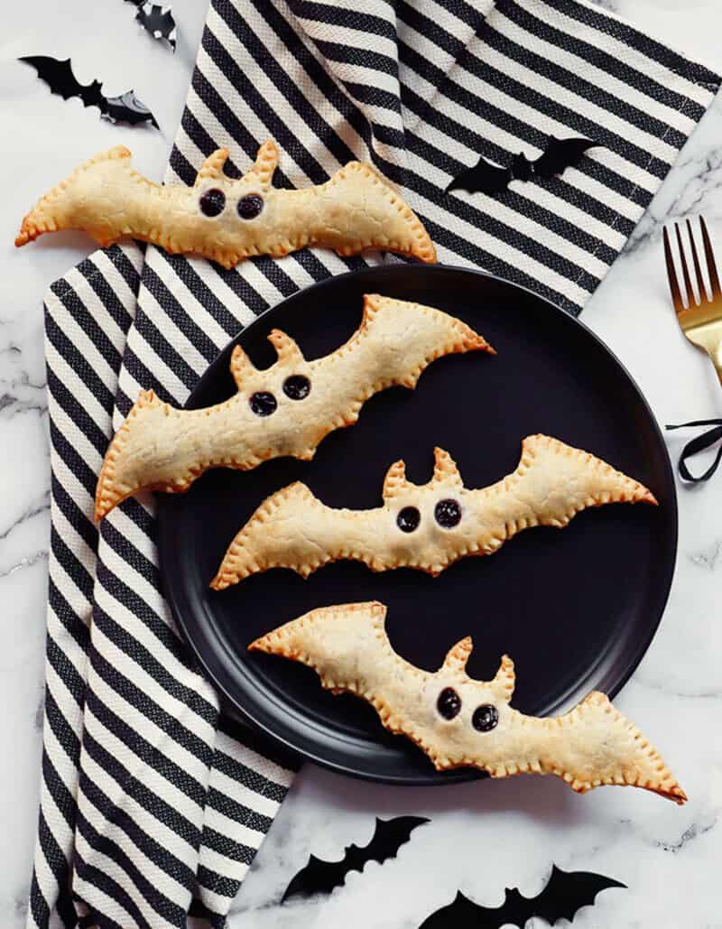 Halloween bat pastries on a black plate with striped napkin and small bat decorations.
