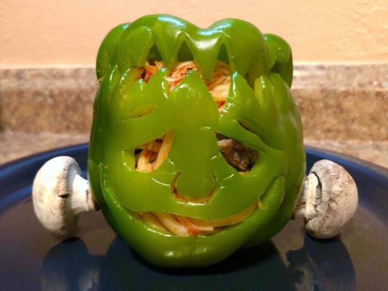 Smiling green bell pepper jack-o'-lantern with mushroom eyes on a blue plate, shredded filling peek.