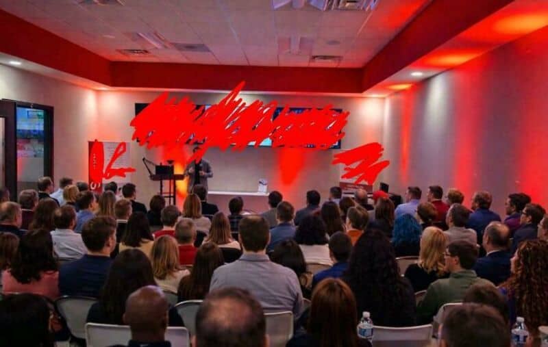 A speaker presenting to a large audience in a conference room with red lighting.