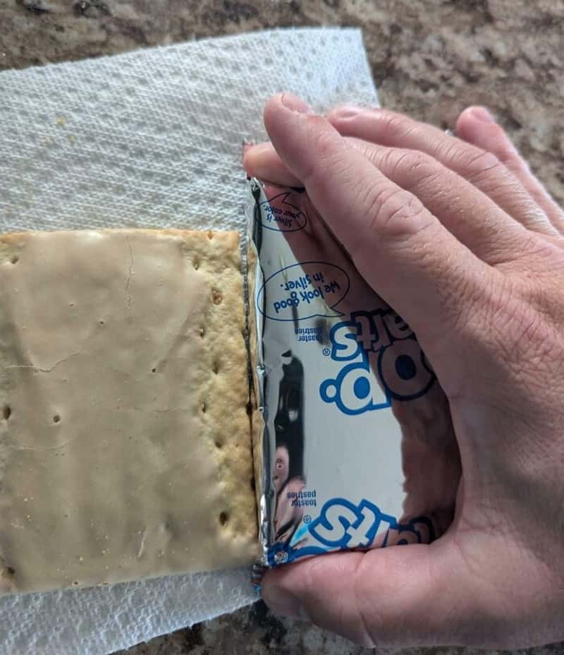 Hand gripping a partially unwrapped rectangular cracker with a beige topping, on a paper towel.