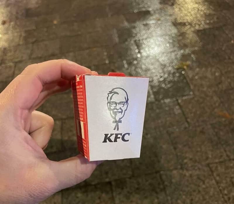 Small KFC-themed box held in a left hand outdoors, with Colonel Sanders logo on front.