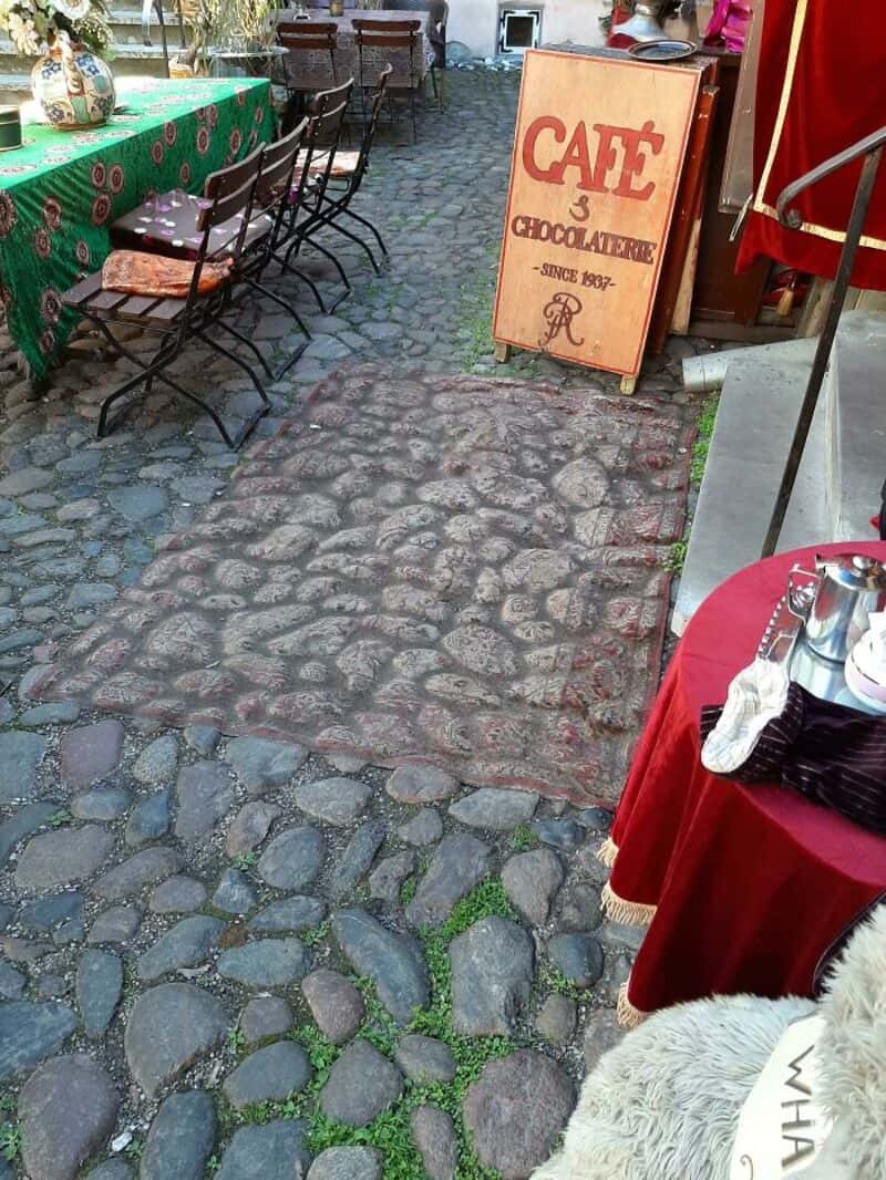 Outdoor cafe with cobblestone path, chairs, and a sign for a chocolate shop.