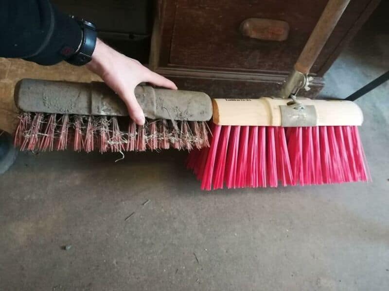 Two brooms with different bristle styles and colors, one worn and one new, on a concrete floor.