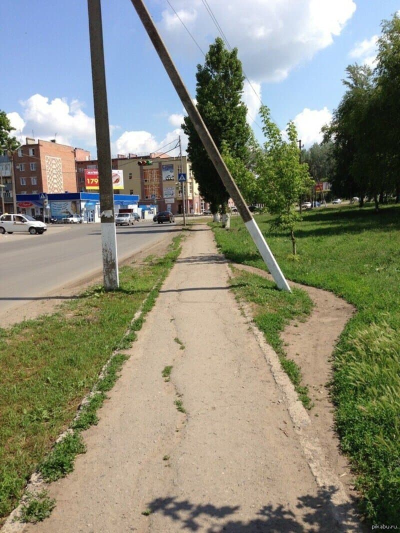 Sidewalk with leaning utility pole, trees, and buildings in the background.
