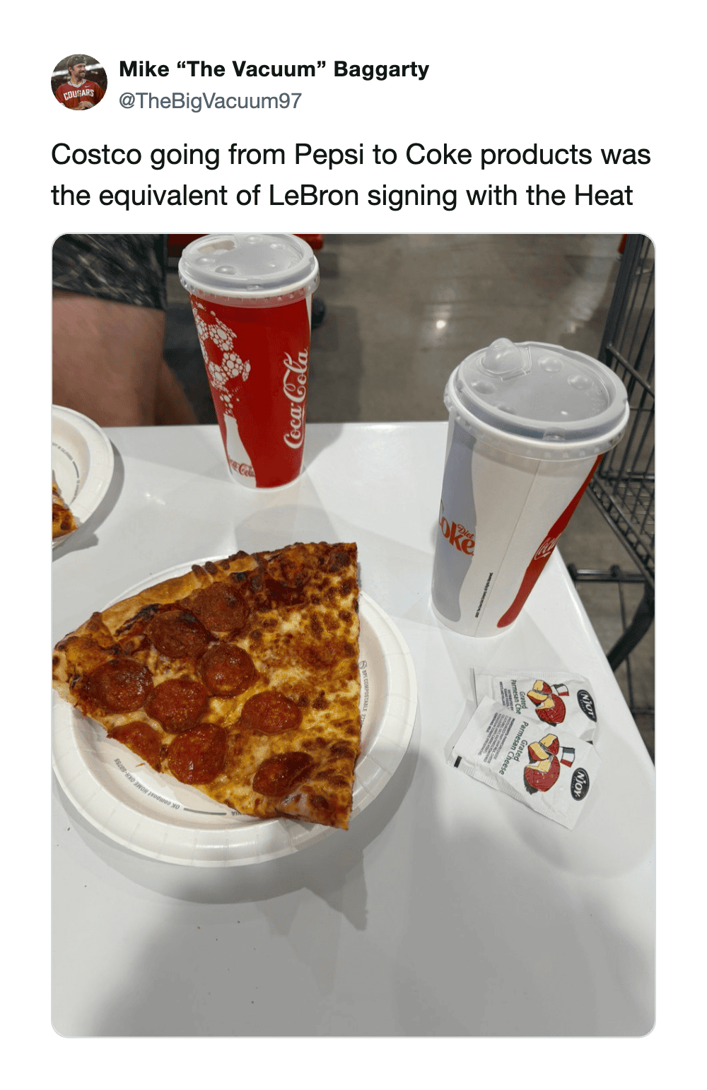 Pizza slice with pepperoni and cheese on a white plate, two soda cups in the background.