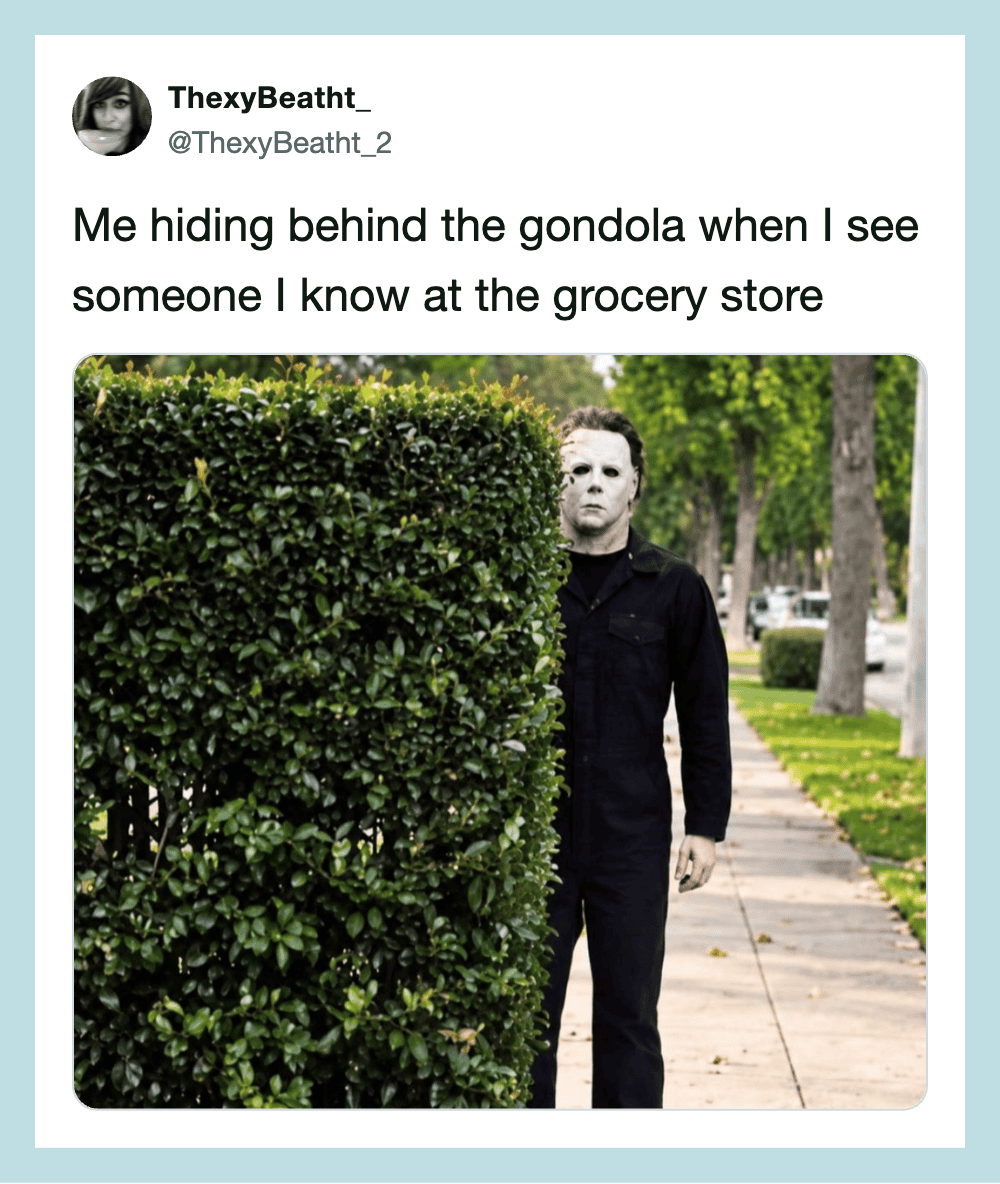 Person hiding behind a large bush on a sidewalk with trees and grass.