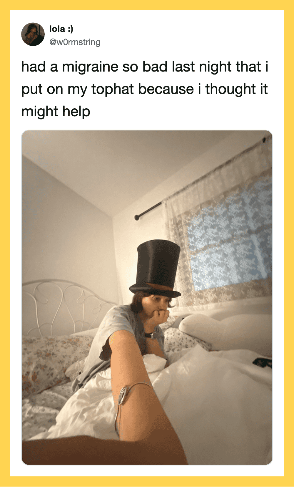 Person wearing a top hat sitting on a bed with a migraine, selfie taken by someone else.