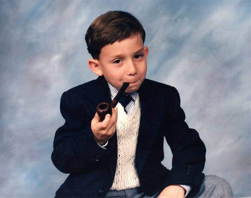 Young boy in a suit smoking a pipe, sitting against a cloudy background.