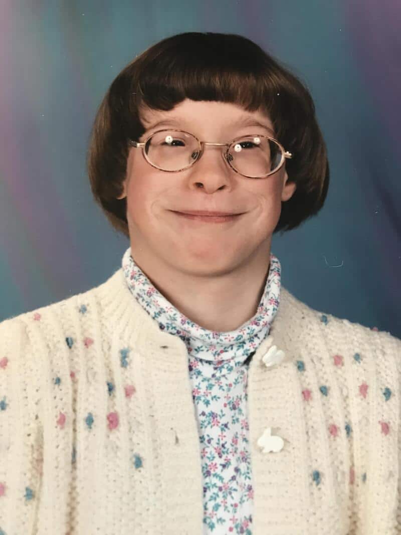Young girl with glasses, short brown hair, smiling, wearing a white cardigan and floral shirt.