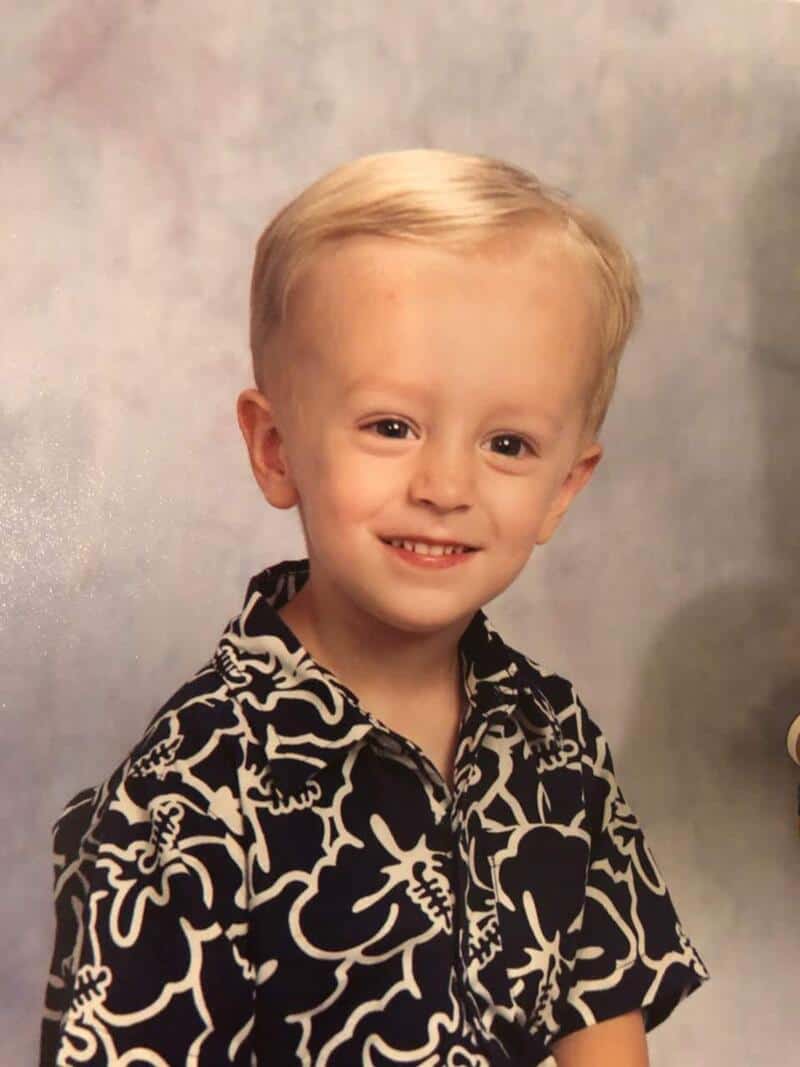Young boy with blonde hair smiling, wearing a black and white patterned shirt.