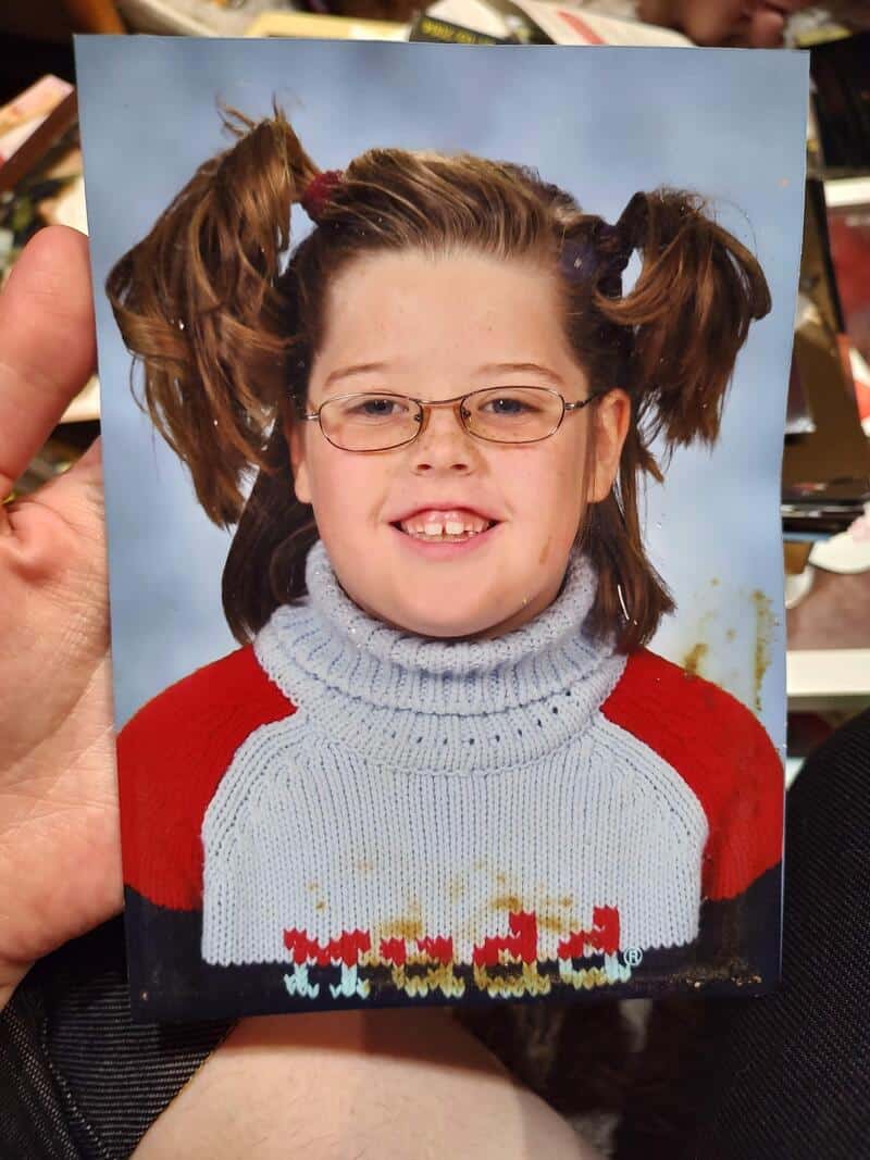 Child with glasses, pigtails, and a colorful sweater, smiling at the camera.