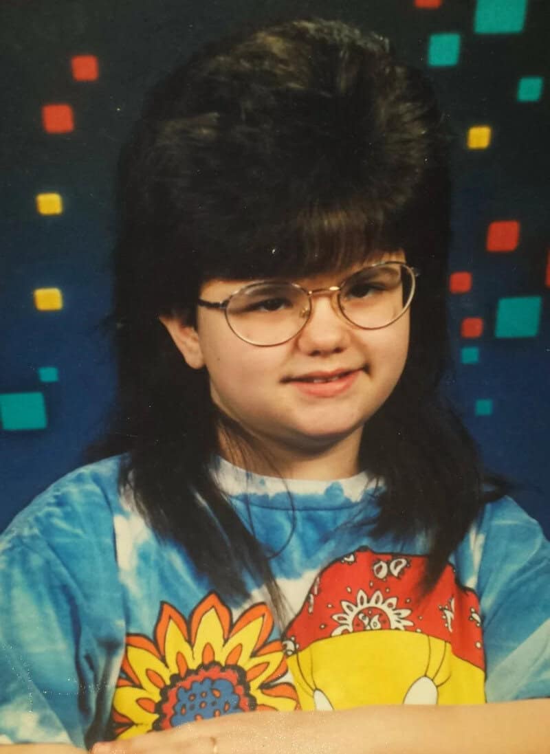 Child with big hair, glasses, and a colorful shirt, smiling in front of a patterned background.