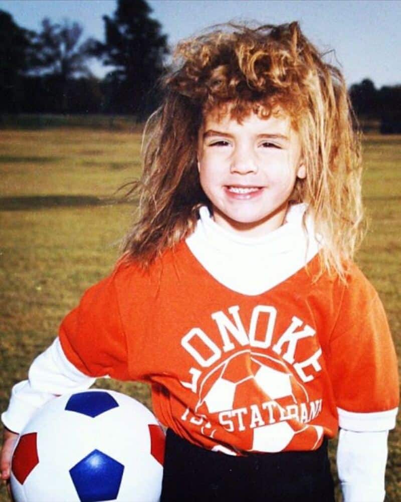 Young girl with curly hair in a soccer uniform holding a soccer ball outdoors.