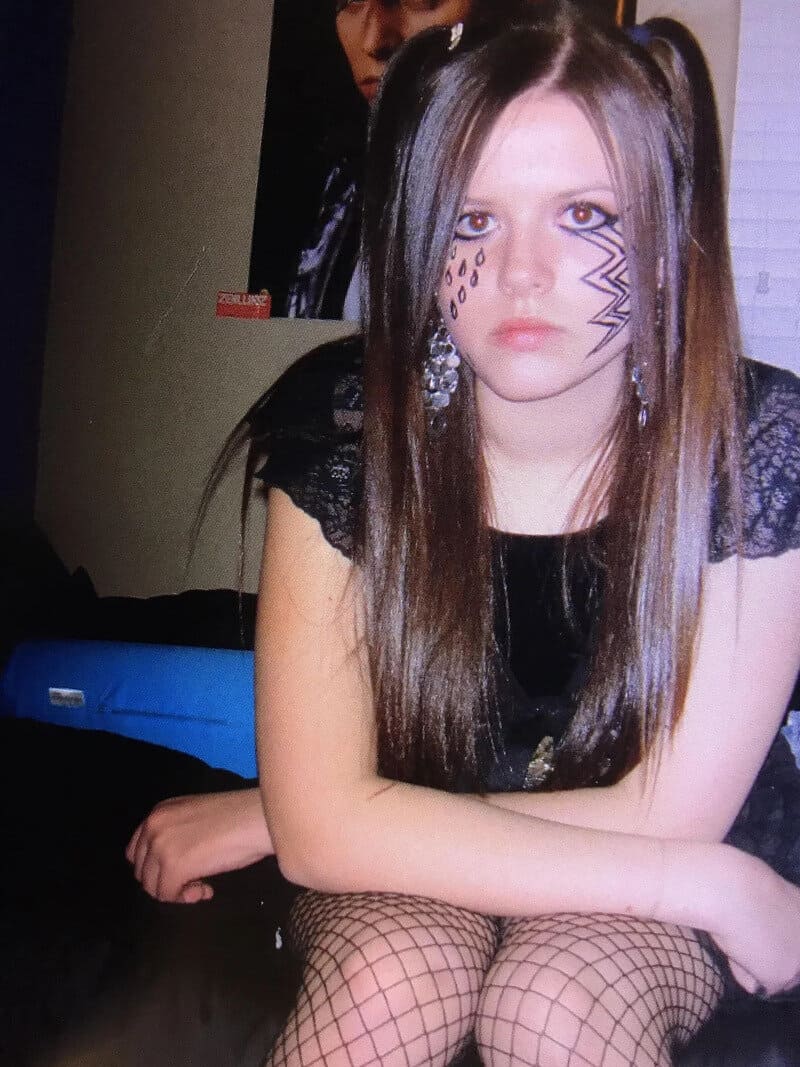 Young woman with face makeup, fishnet stockings, and earrings, sitting with arms crossed.