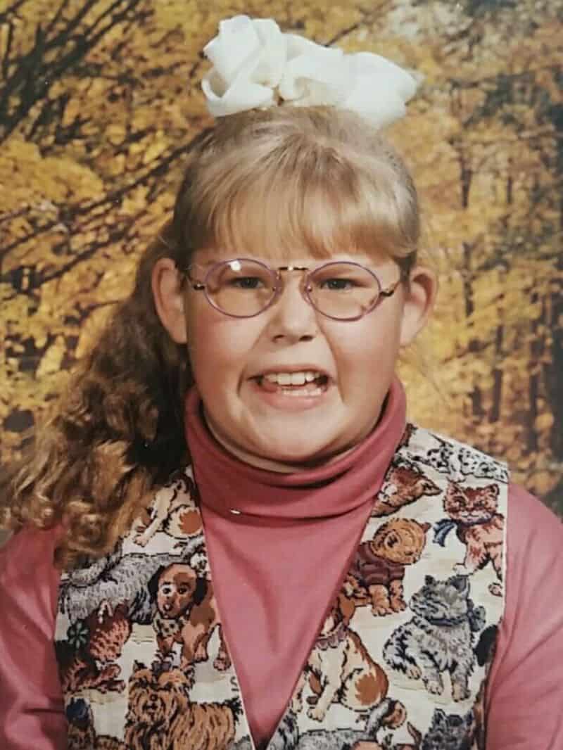 Young girl with glasses, a white bow, and a tiger-patterned vest, smiling.