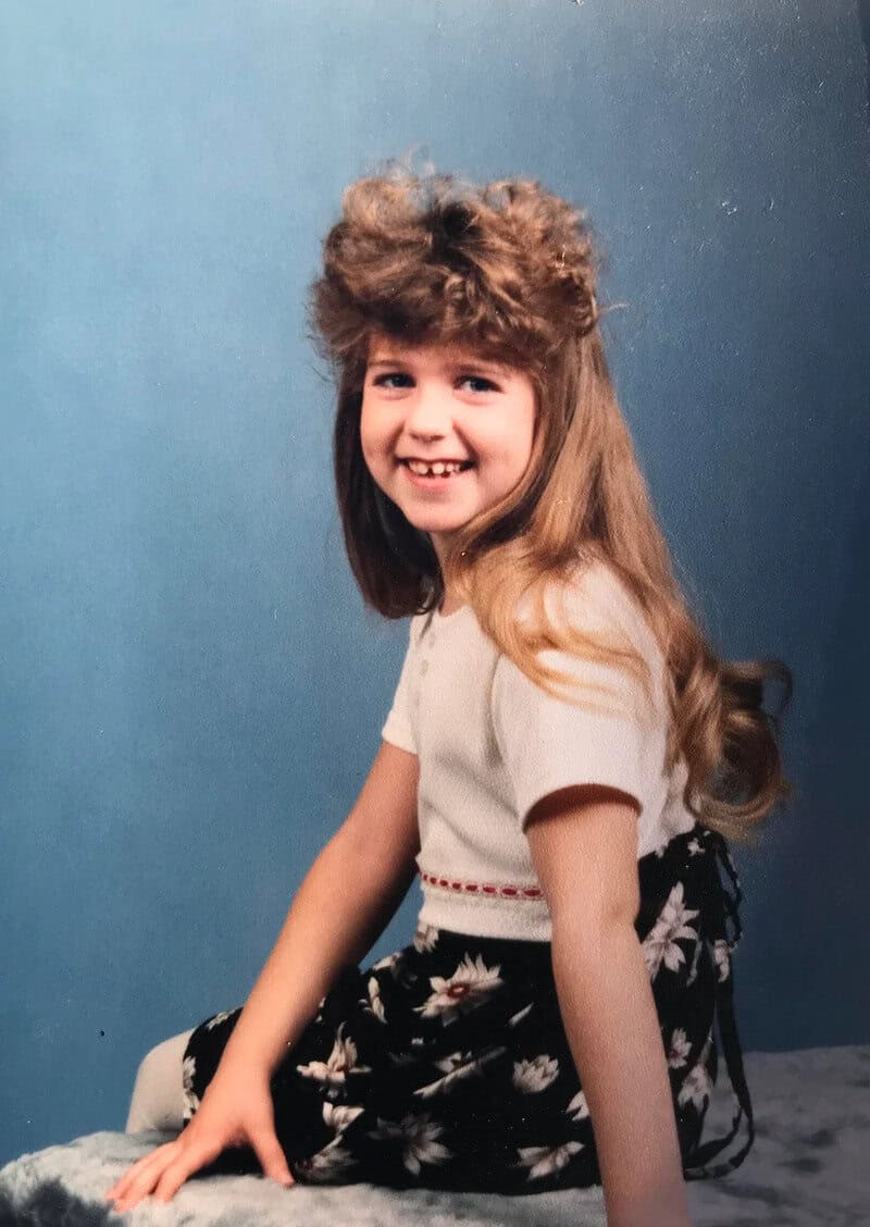 Young girl with long hair and a big curly hairdo, smiling, sitting on a chair.