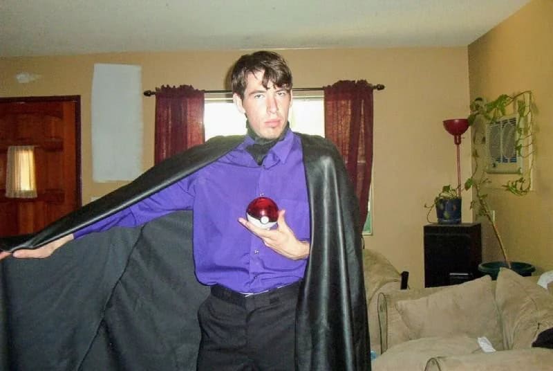 Man in a purple shirt with a black cape holding a red ball in a living room.