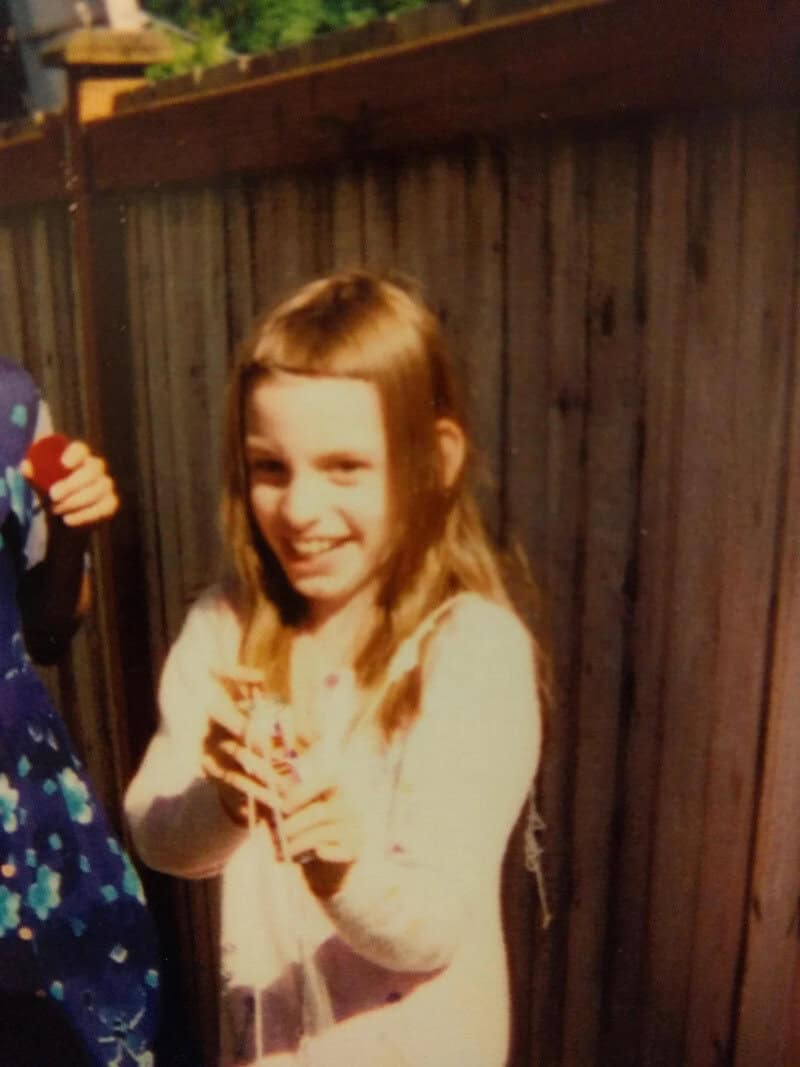 Smiling girl with long hair holding a small object, standing near a wooden fence.