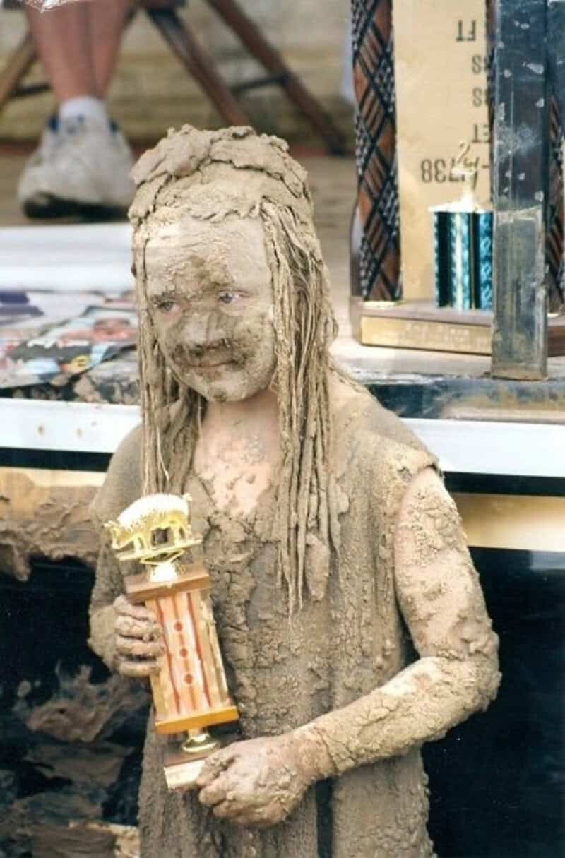 Child covered in mud holding a trophy, smiling, with books and people in the background.