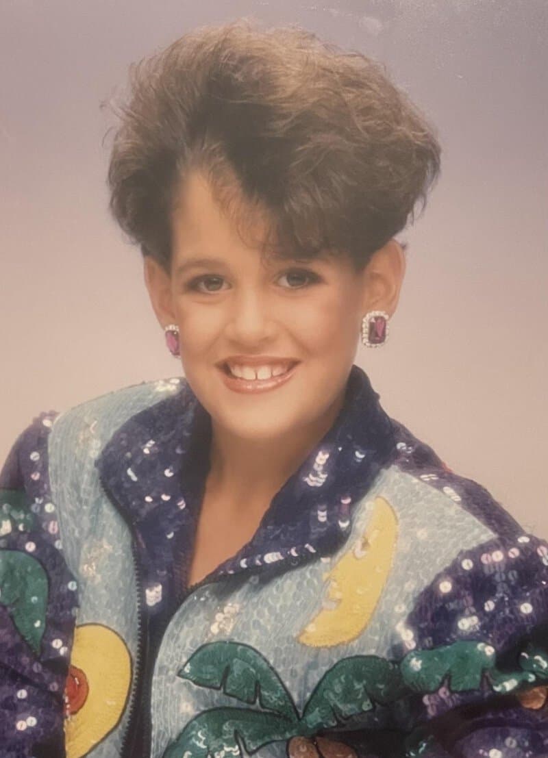 Young woman with big hair, colorful jacket, and earrings, smiling at the camera.