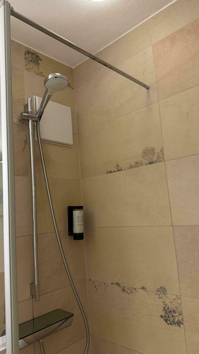 Shower with a soap dispenser and a shower head on a beige tiled wall.