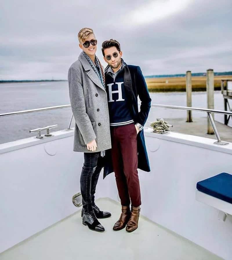 Two men in stylish outfits posing on a boat deck near water with a railing and pier in the distance.