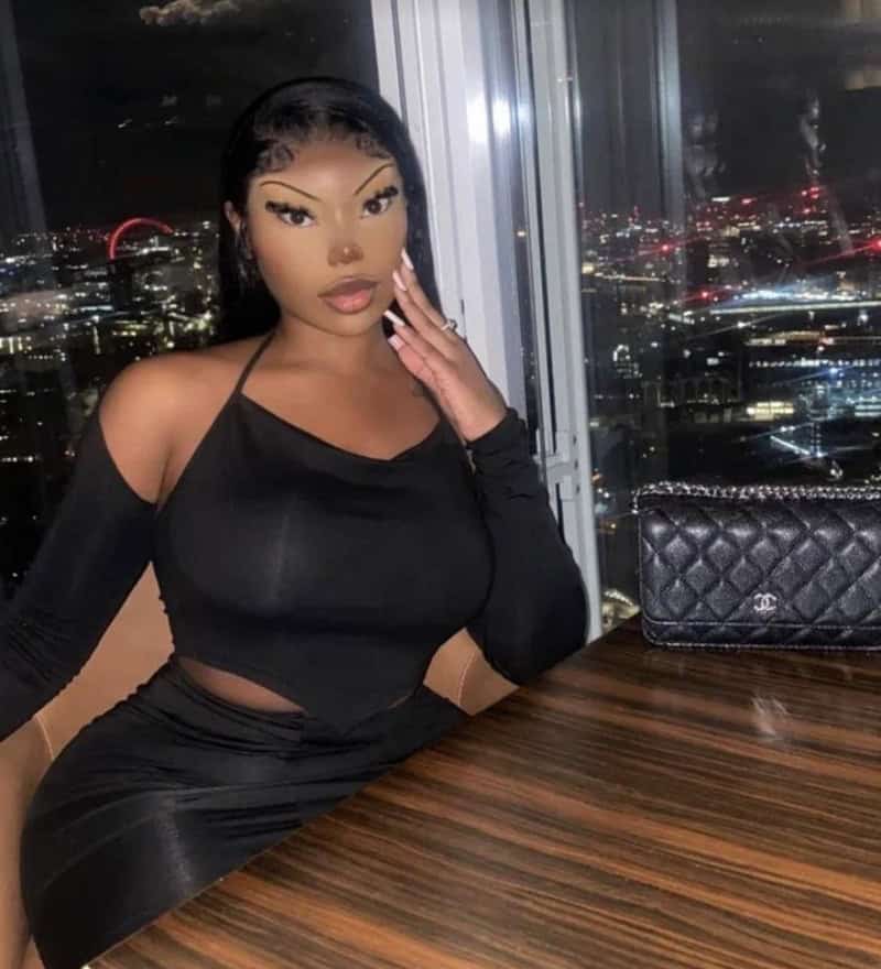 Woman in black off-shoulder top at night with city lights through window.