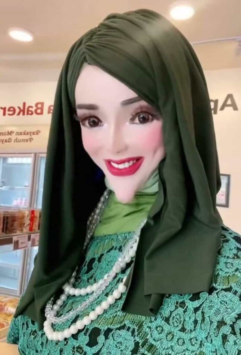 Portrait of a woman wearing a dark green hijab and matching lace top, with a pearl necklace and pink.