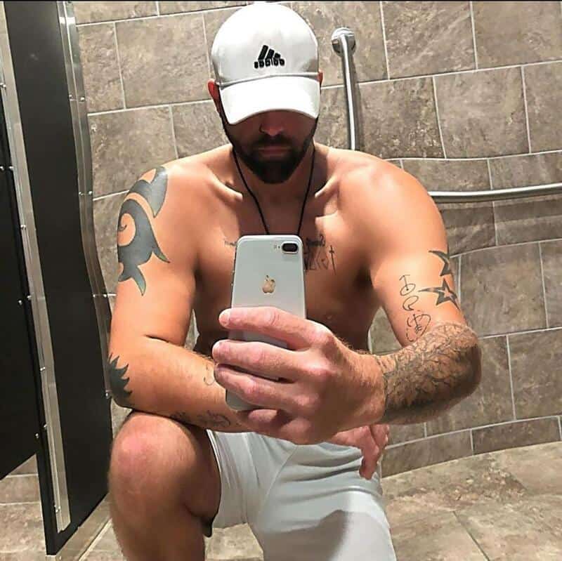 Man in a white cap shirtless in a tiled bathroom taking a mirror selfie with his phone.
