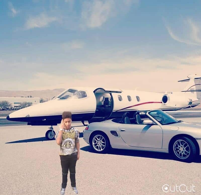 Young boy standing in front of a white private jet and a silver convertible car on a tarmac.
