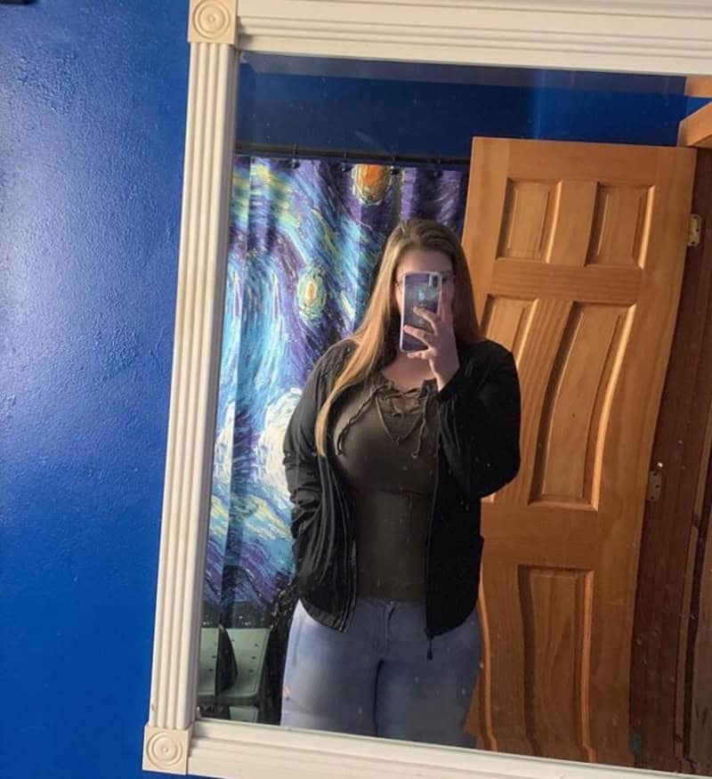 Woman taking a mirror selfie in a blue-walled room, wearing a black jacket over a green top and blue.