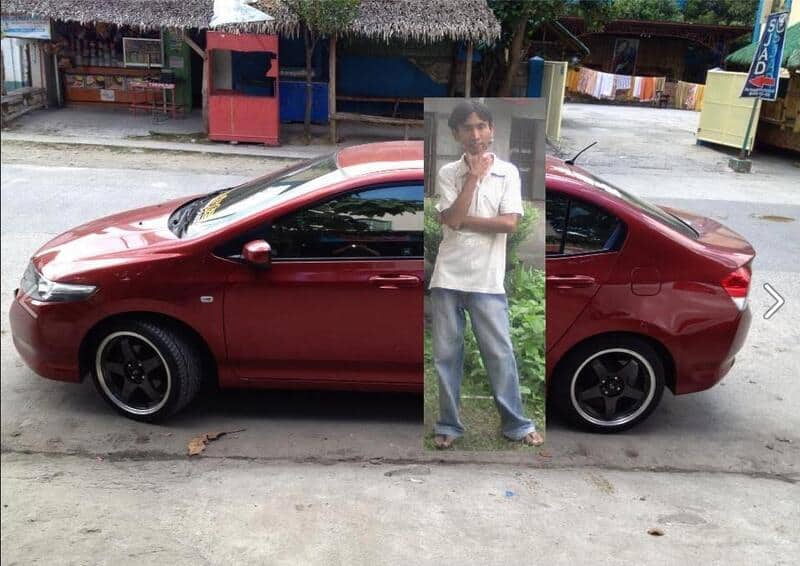 A man standing in front of a red sedan on a street with shops in the background.