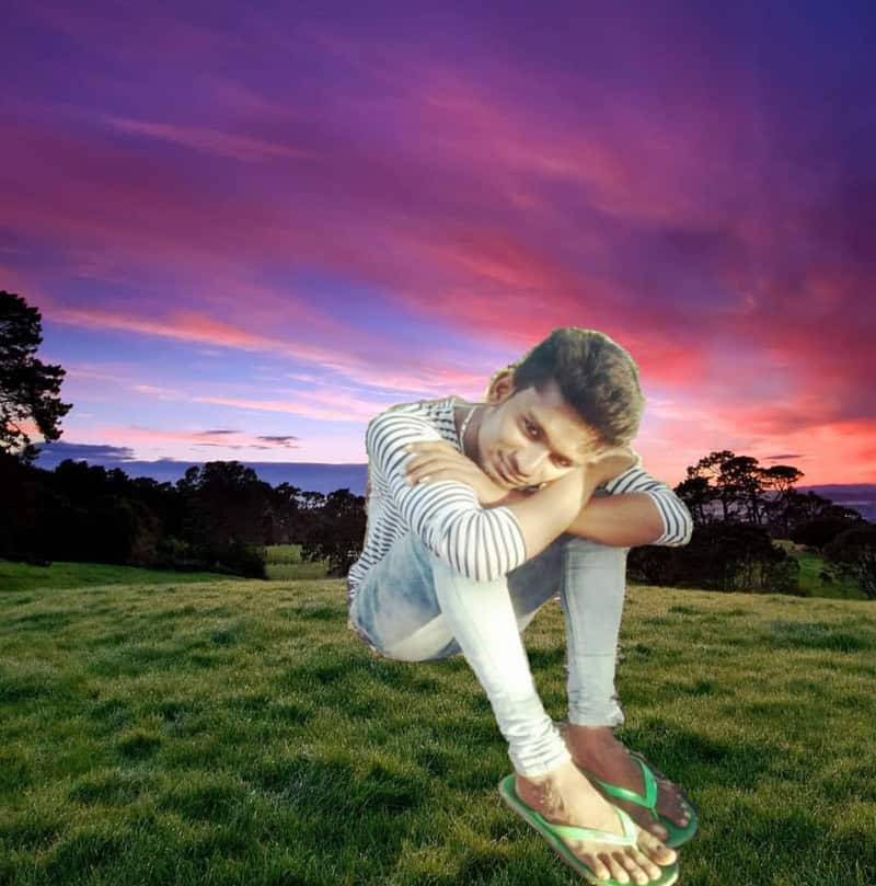 Young man sitting on grass at sunset wearing striped shirt and jeans with green flip-flops, headrest.