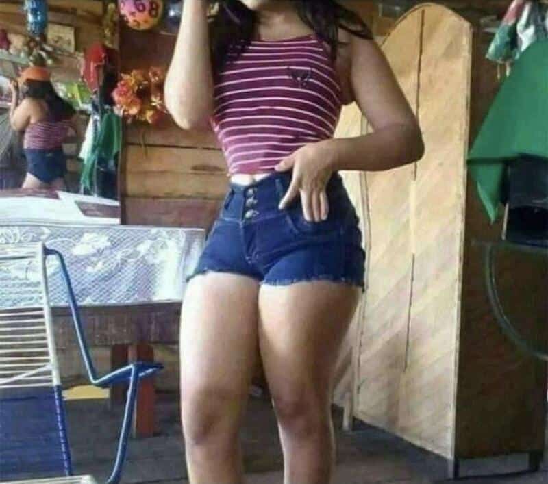 Young woman in striped crop top and high-waisted denim shorts standing indoors; blurred background.